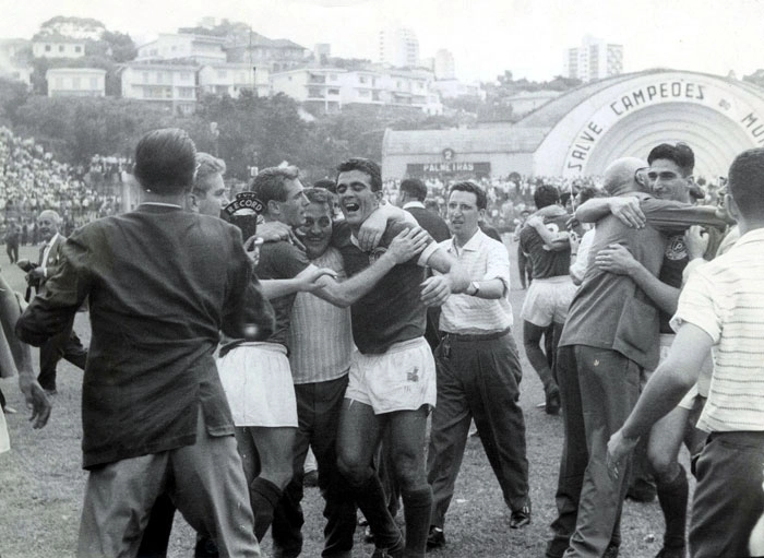 No dia 10 de janeiro de 1960, o Palmeiras sagrou-se campeão campeão paulista de 1959, em uma vitória sobre o Santos, por 2 a 1, no estádio do Pacaembu. A foto maravilhosa mostra o excepcional repórter Reali Júnior, com o microfone da Rádio Record, tentando entrevistar Geraldo Scotto, um dirigente palmeirense, e o ex-ponta Nardo. Do lado direito, você um dos maiores atacantes da história do futebol brasileiro em todos os tempos, Julinho Botelho, abraçando um senhor calvo.  O palmeirense de costas, sendo abraçado por muitos, é o zagueiro Waldemar Carabina, com a camisa número 3. Atrás deles, Américo Murolo. Ao fundo, ao lado da concha acústica, é possível ler o nome Palmeiras no placar e o número 2 acima das letras