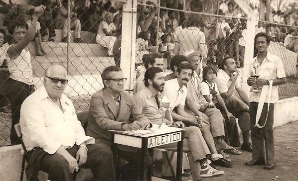 Os torcedores estão na arquibancada. À beira do gramado, da esquerda para a direita: Nilo Bortoloti, José Octaviano Sales, Joaquim Pampa, Vander Magalhães (parcialmente encoberto), Michel Salomão (de bigode) e o último sentado, com a mão no queixo, é Amintas Inacarato. À direita, em pé, está Vitor Padeiro. Atrás, com a mão no alambrado está Zé Carlos Bonaldi. Foto enviada por Celio Sales