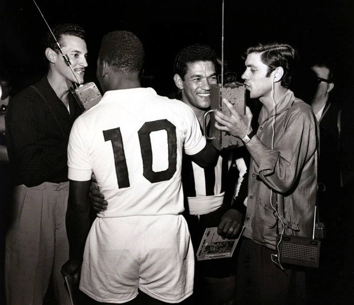 Veja que foto fantástica em um clássico Santos x Botafogo, provavelmente fim da década de 50 e início de 60, dois gênios do futebol: Pelé e Garrincha, respectivamente defendendo Santos e Botafogo e veja, os grandes repórteres, à esquerda Tom Barbosa da Rádio Record e Luis Aguiar, da Rádio Bandeirantes. Notem seus equipamentos radiofônicos, que diferença dos celulares de hoje, tão frequente nas transmissões esportivas. Foto In my Ear