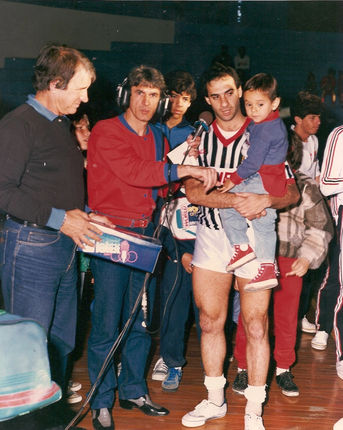 Da esquerda para a direita, Pisco Palumbo (o segundo) com microfone em punho entrevista o ex-craque do futsal Douglas, que segura criança no colo. Foto: Sarkis