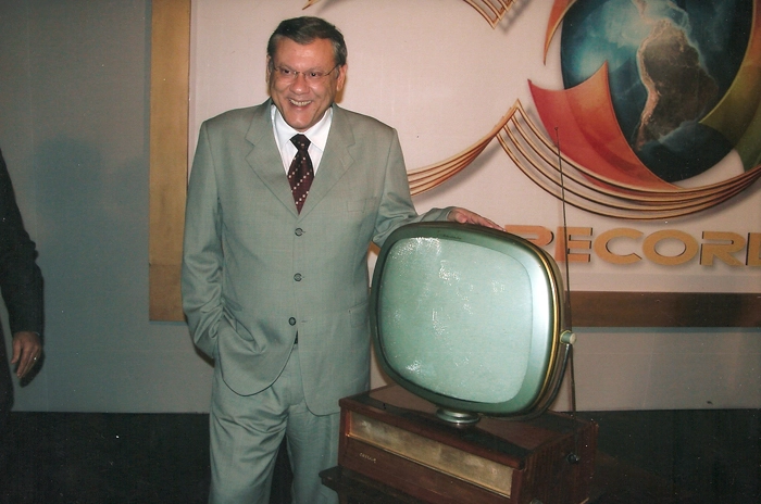 Milton Neves fica deslumbrado com o antigo aparelho de TV que está ao seu lado. Foto cedida pela Rede Record 