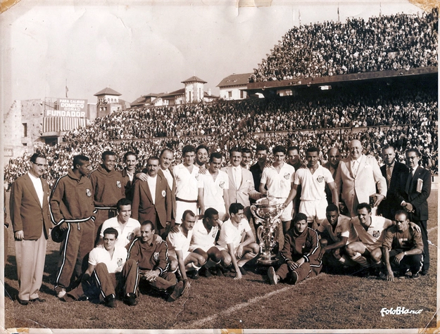 A foto, do dia 21 de junho de 1959, foi tirada na cidade espanhola de La Coru&ntilde;a. Maravilhosa por si s&oacute;, mostra o Peixe posando 30 minutos depois de um jogo em que o goleou o Botafogo por 4 a 1 e ficou com a ta&ccedil;a do torneio Tereza Herrera, um dos mais tradicionais do mundo disputado desde 1946. Por sinal, o alvinegro da Vila jogou tanto naquela ocasi&atilde;o que a torcida n&atilde;o arredou p&eacute; do est&aacute;dio, l&aacute; ficando para aplaudir ininterruptamente os craques que bailavam com a camisa branca mais famosa do mundo. Em p&eacute; est&atilde;o o tesoureiro Ciro Costa, Dorval, Fioti, o inesquec&iacute;vel Fiori Giglioti, o m&eacute;dico da delega&ccedil;&atilde;o Dal&oacute; Salerno, Lula, Get&uacute;lio, cartola n&atilde;o identificado, Zito, Athi&eacute; Jorge Cury, Formiga, Lal&aacute;, Ramiro, La&eacute;rcio, Mour&atilde;o, mais um cartola n&atilde;o identificado, Modesto Roma e mais dois membros da delega&ccedil;&atilde;o n&atilde;o identificados. Na fila de baixo est&atilde;o Feij&oacute;, Dalmo, Pav&atilde;o, Alfredinho, Coutinho, &Aacute;lvaro, Afonsinho, Pel&eacute;, Pepe e o massagista Macedo com a camisa com a letra 