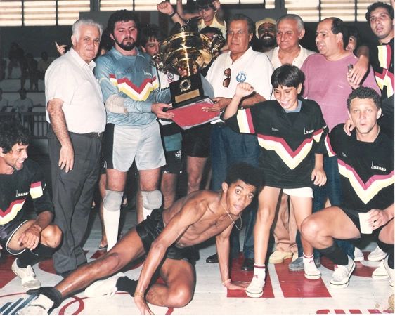 De camiseta branca e óculos pendurado, o ex-presidente do Corinthians, Alberto Dualib, carrega o troféu conquistado no futebol de salão. Reparem no atleta, que aparece abaixado, só de bermuda, se esforçando para conseguir aparecer na foto. Foto: Sarkis