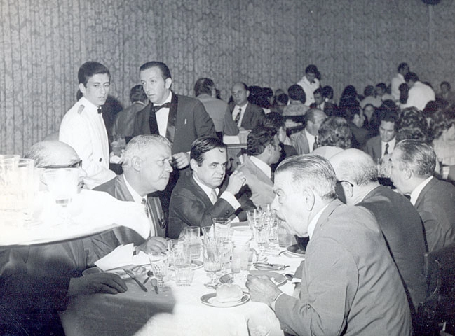 No lado esquerdo da mesa, da esquerda para a direita: Ary Silva, Mauro Pinheiro, Roberto Silva, e ao fundo, na ponta mesa, aparece Joseval Peixoto que conversa com Ênnio Rodrigues (que está a sua direita)